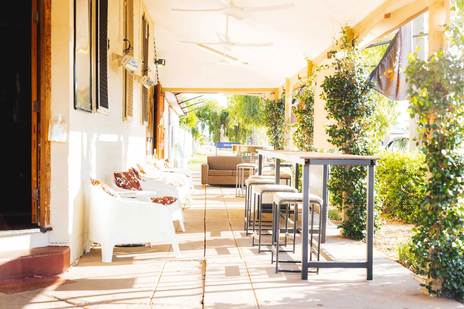 Sunlit verandah at Club Boutique Hotel Cunnamulla with white tub chairs, bar stools and climbing greenery